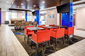 A modern hotel lobby with a mix of seating options, including red upholstered chairs and tables, a patterned carpet, and a large flat-screen TV mounted on a blue pillar.