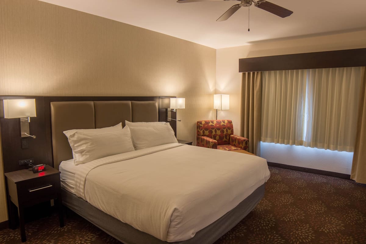 A hotel room with a large bed, two bedside lamps, a nightstand, a chair, and a window with curtains.