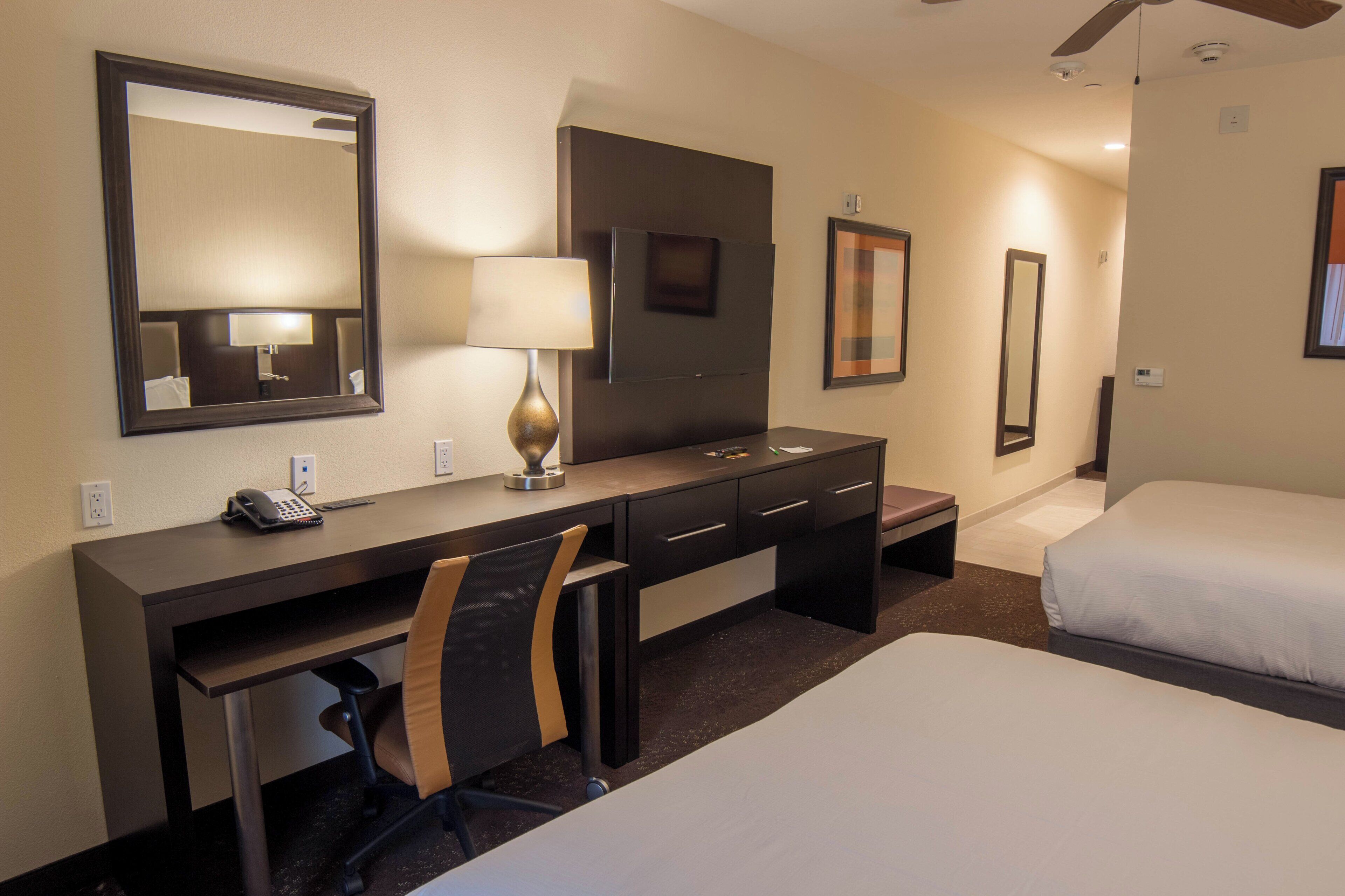 A hotel room with a desk, chair, two beds, a lamp, and a mirror.