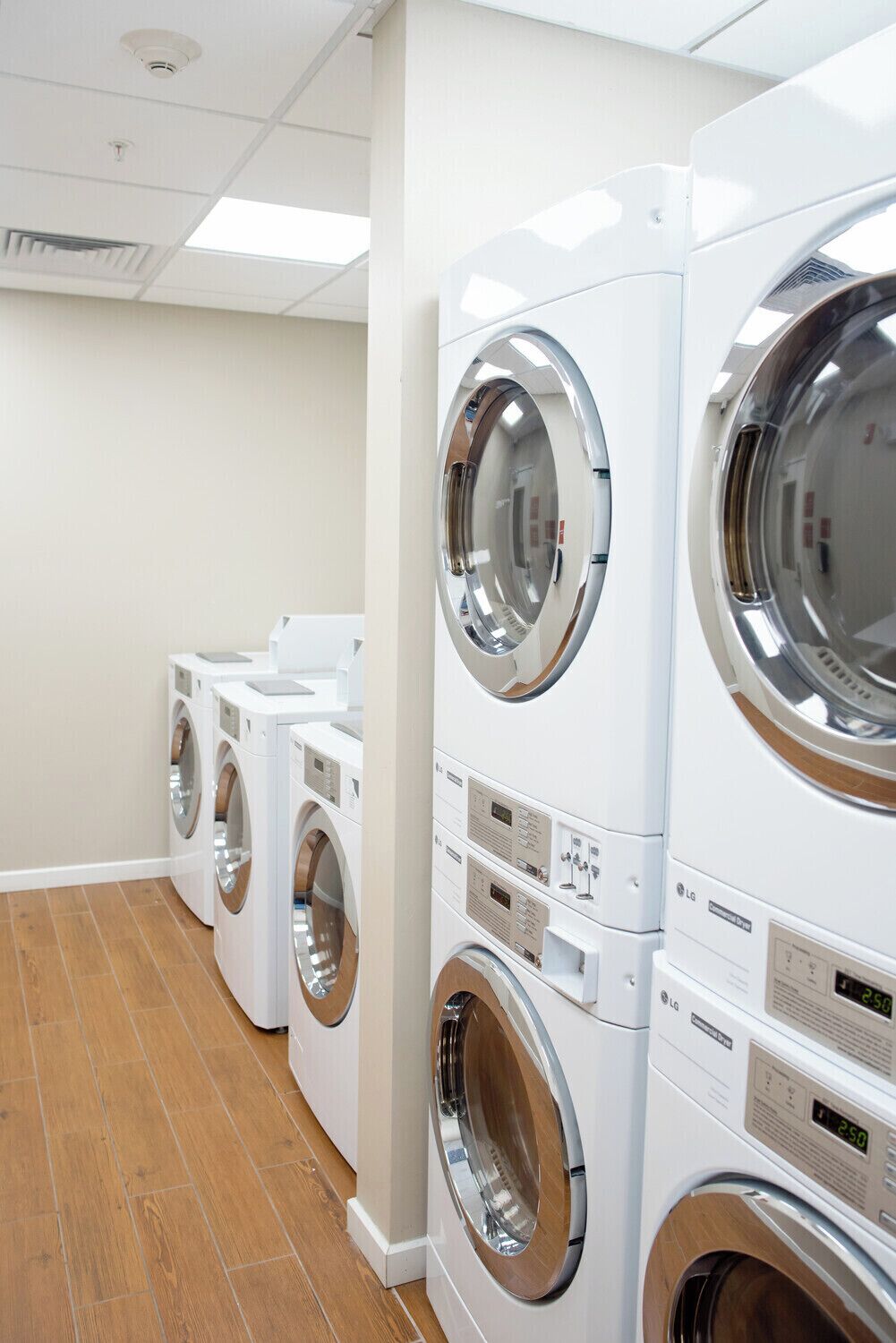 A row of white front-loading washers in a laundry room with wooden flooring and white walls.