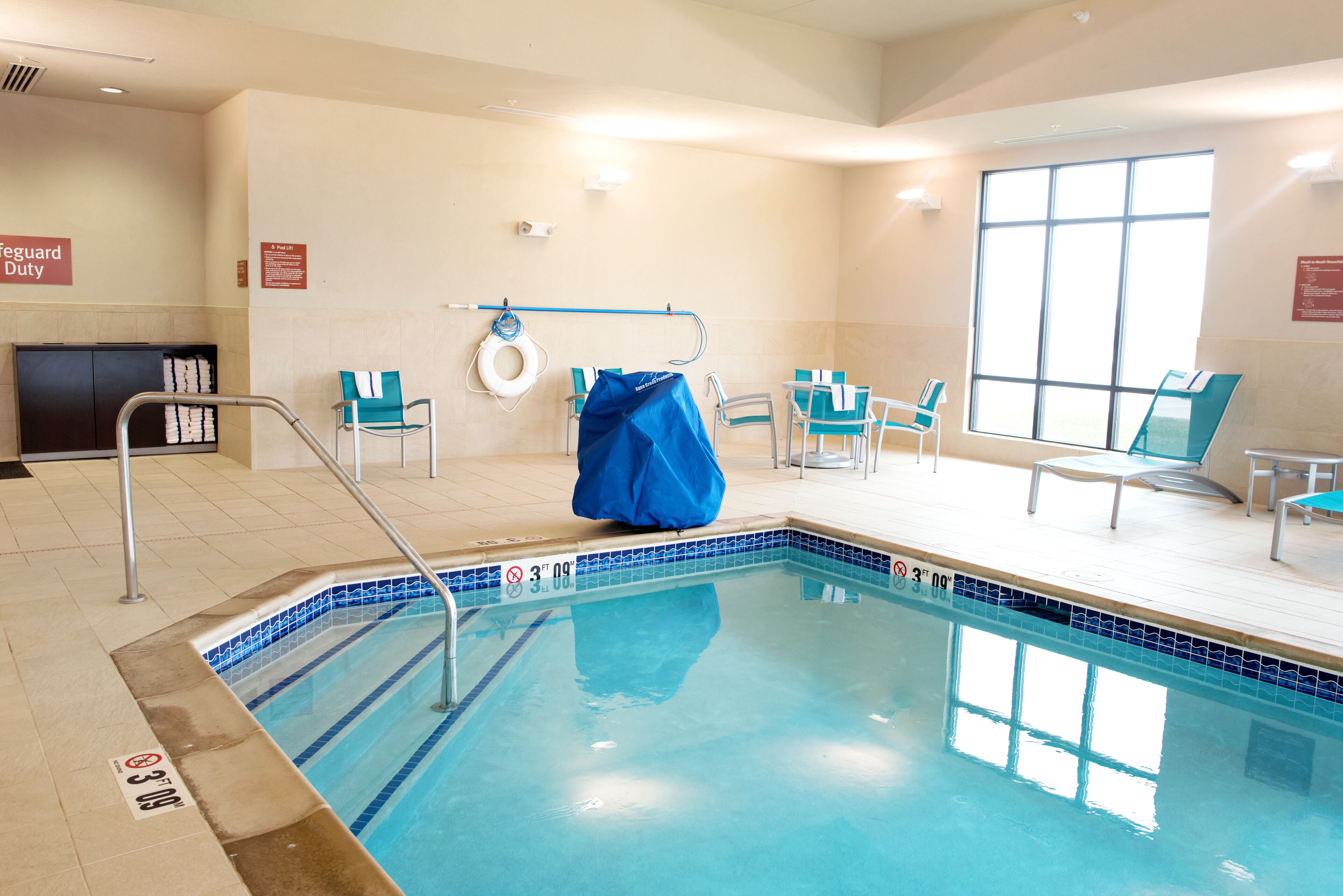 An indoor swimming pool with clear water, a lifeguard duty sign, and a blue cover over the pool.