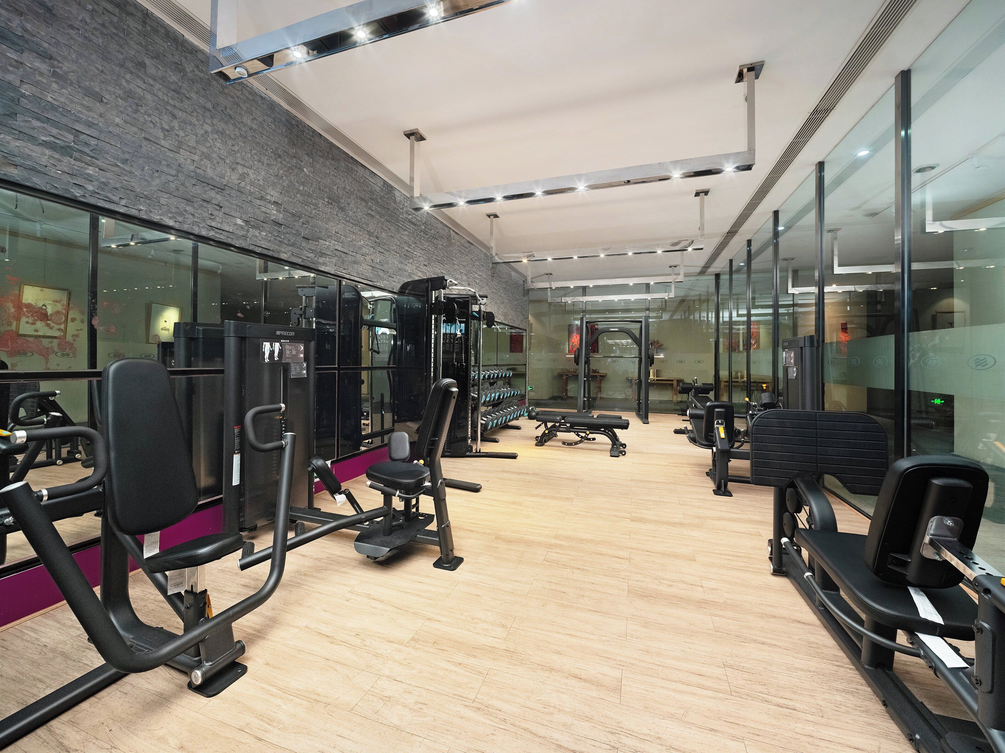 A modern gym with various exercise equipment, glass walls, and a wooden floor.