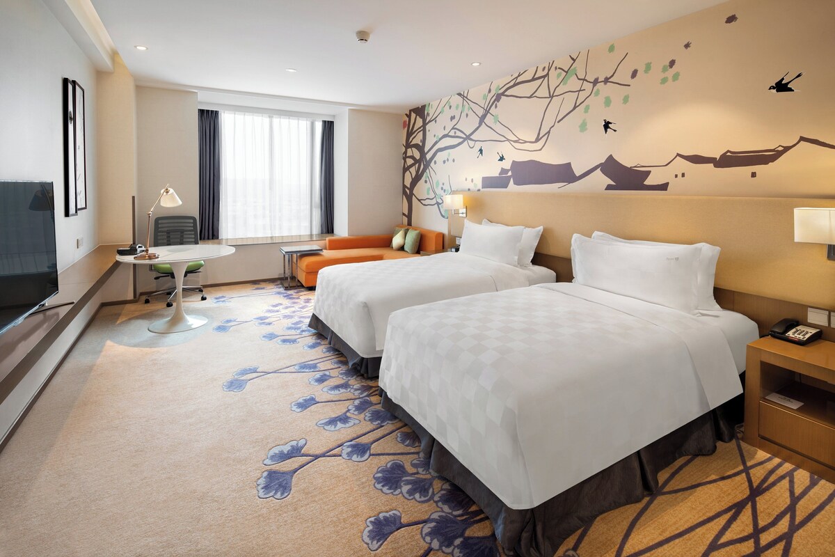 A hotel room with two beds, a desk with a chair, a sofa, a TV, and a large window.