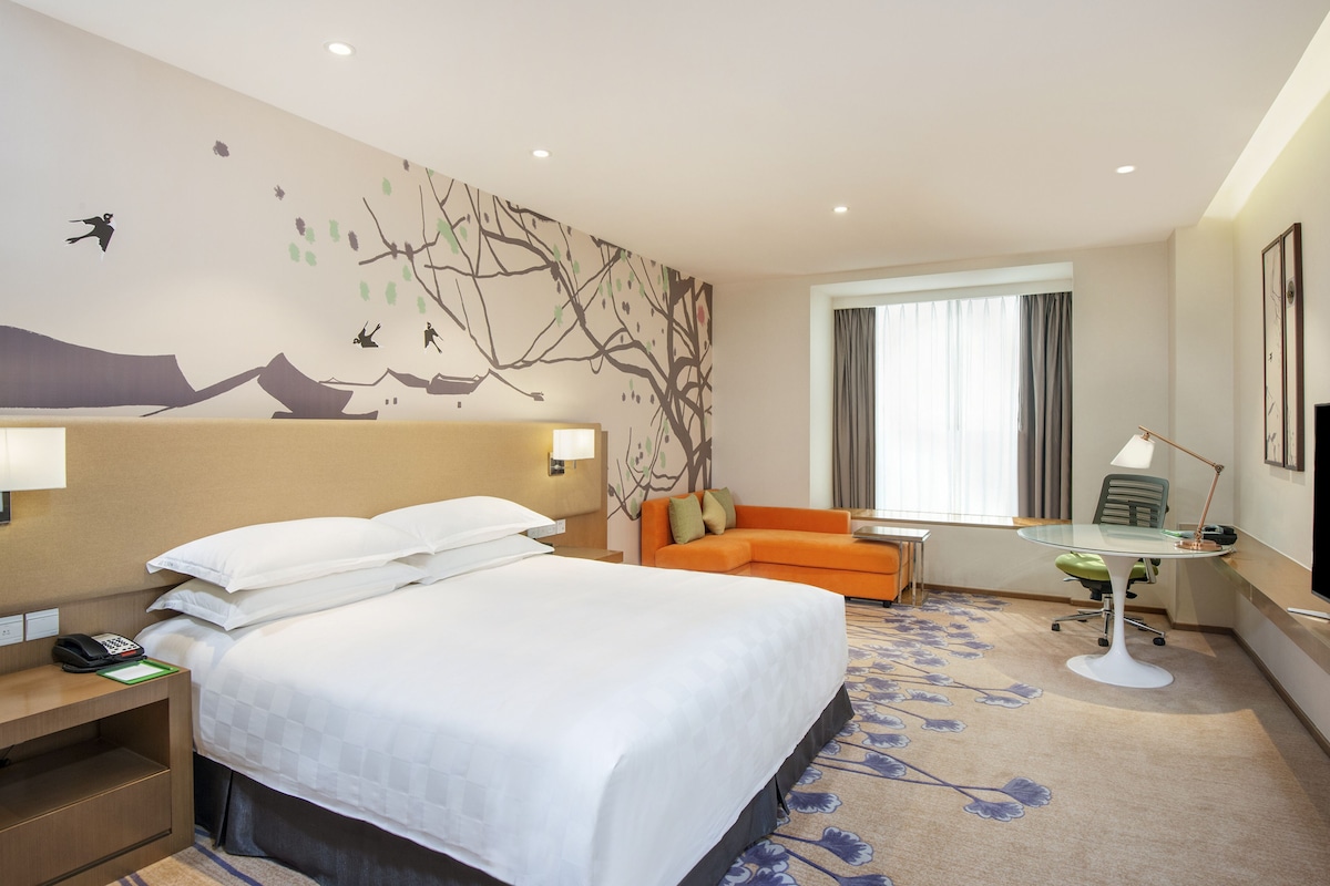 A hotel room with a large bed, a sofa, a desk with a lamp, and a window with curtains.