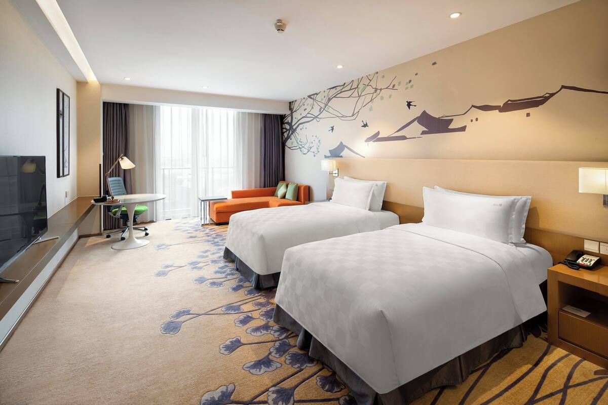 A hotel room with two beds, a desk with a computer, a TV, and a large window with curtains.