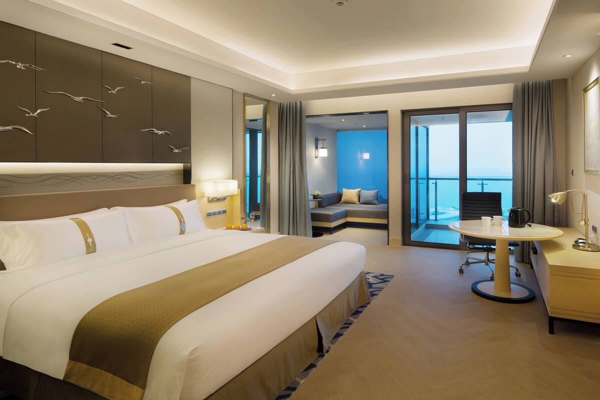 A hotel room with a large bed, a desk with a chair, a small table, a TV, and a balcony with a view.