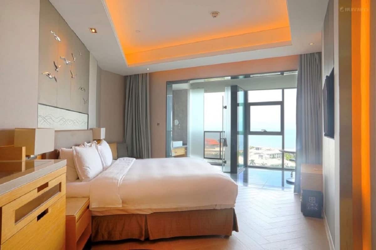 A modern hotel room with a large bed, a wooden dresser, a glass door leading to a balcony with a view, and a ceiling with recessed lighting.