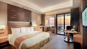 A hotel room with a large bed, a desk, a chair, a TV, and a balcony with a view.
