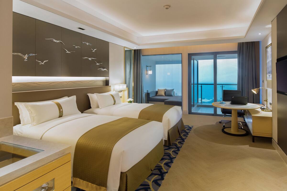A hotel room with two beds, a desk with a laptop, a TV, and a balcony with a view of the sea.