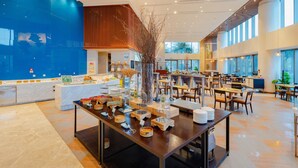 Un vaste espace dédié à la salle à manger, avec de nombreuses tables et chaises, une plante décorative au centre, et un design moderne agrémenté de grandes fenêtres et d’un mur d’accent bleu.