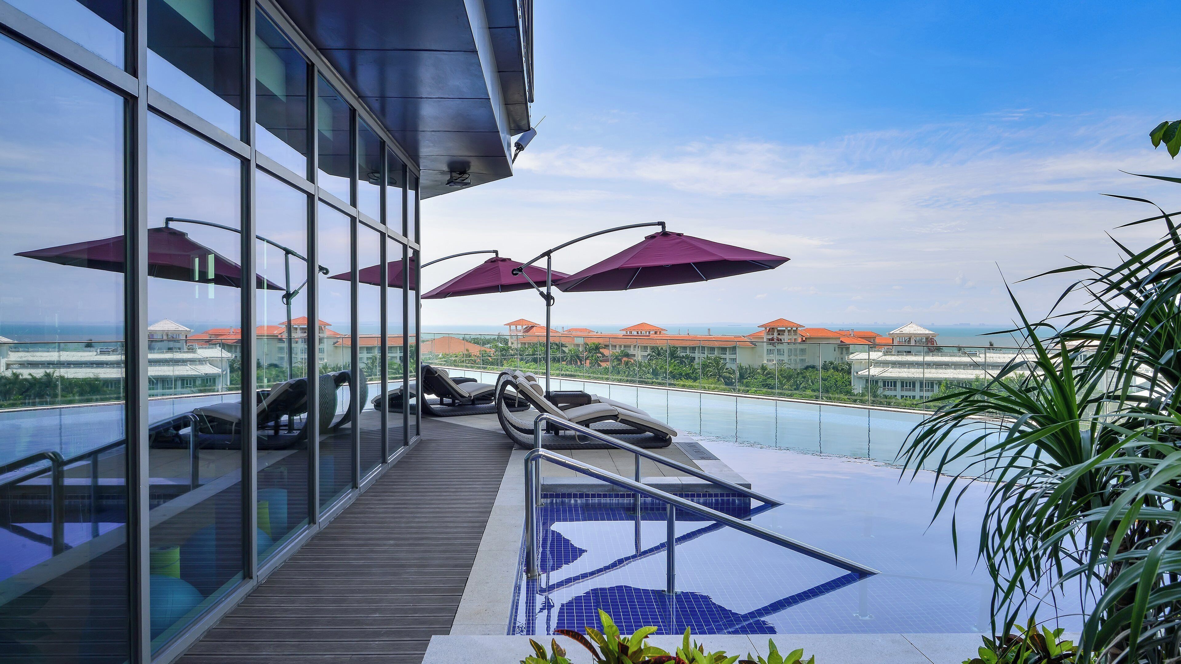 A rooftop pool area with lounge chairs and umbrellas, overlooking a resort with buildings and a beach.