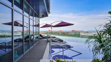 A rooftop pool area with lounge chairs and umbrellas, overlooking a resort with buildings and a beach.