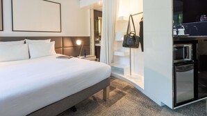 A modern hotel room with a large bed, a bedside table with a lamp, a wardrobe, and a bathroom visible through a glass door. - Novotel Suites Paris Expo Porte de Versailles (Paris)