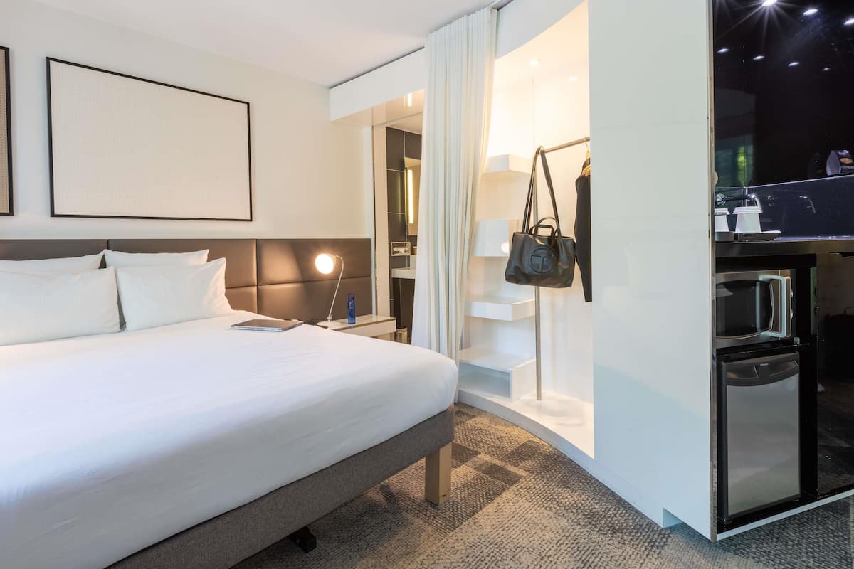 A modern hotel room with a large bed, a bedside table with a lamp, a wardrobe, and a bathroom visible through a glass door.