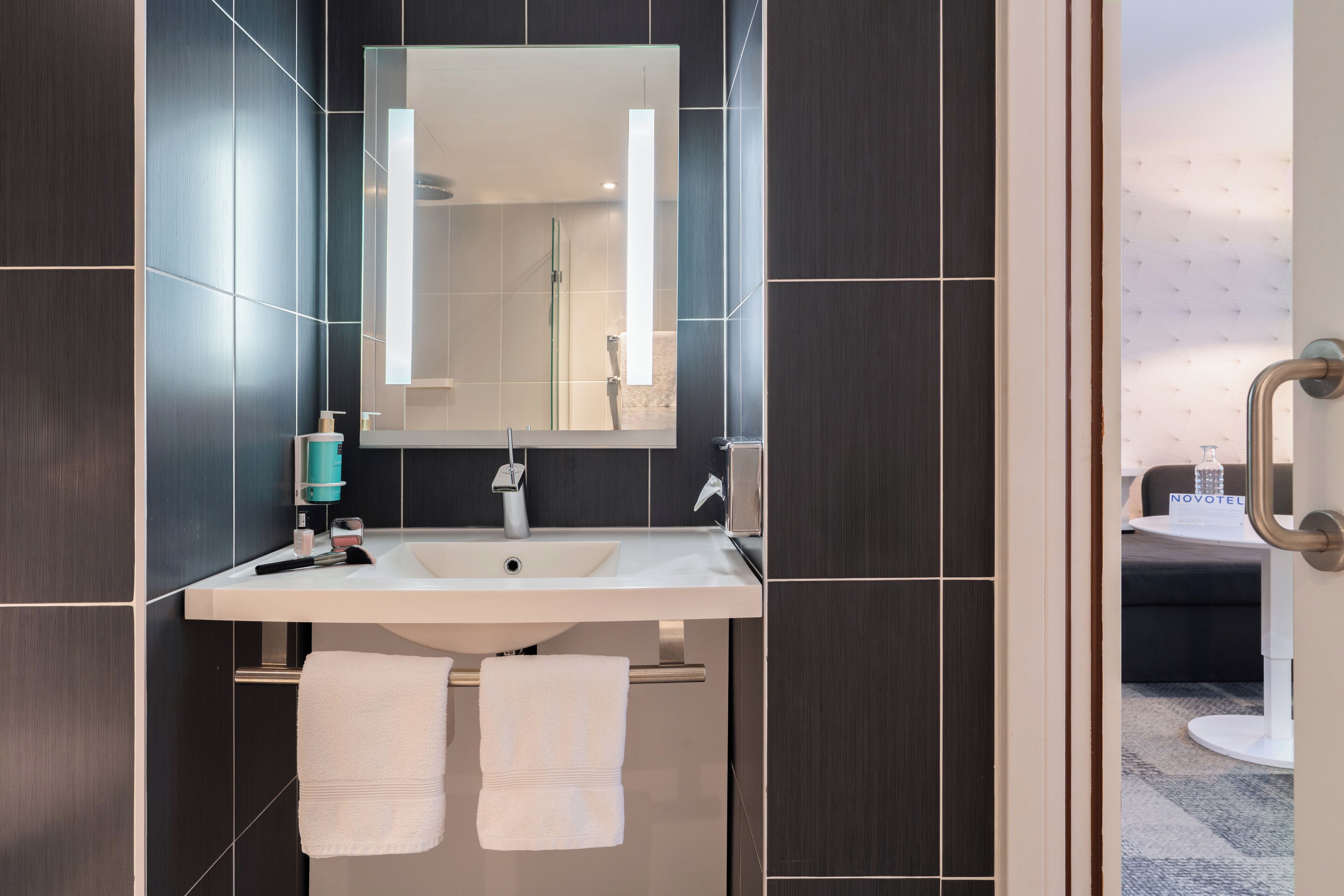 A modern bathroom with dark tiled walls, a white sink, a large mirror, and two white towels hanging on a rack.