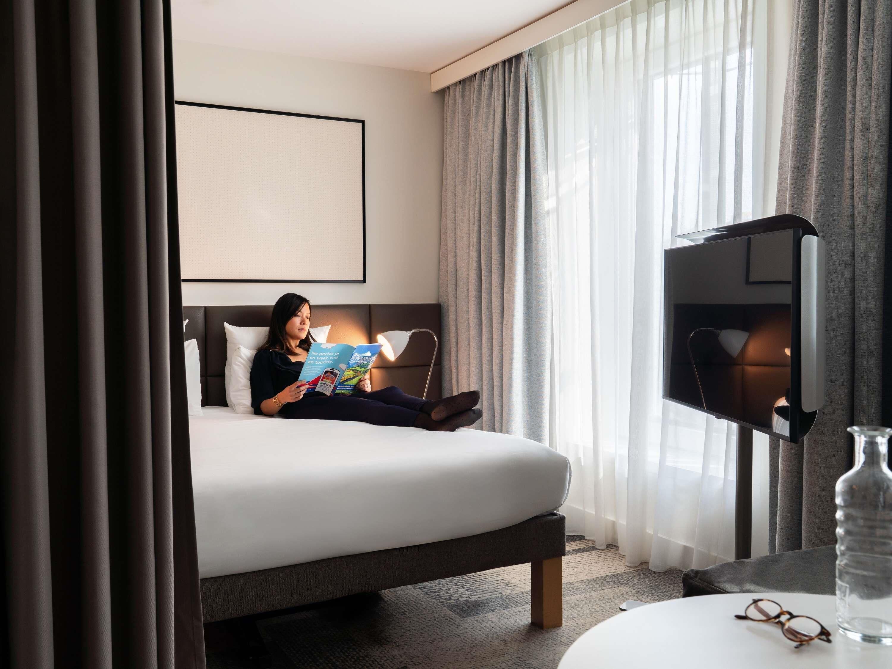 Une chambre d’hôtel avec un lit, un téléviseur à écran plat, une lampe de lecture, une bouteille en verre et une paire de lunettes posées sur une table.