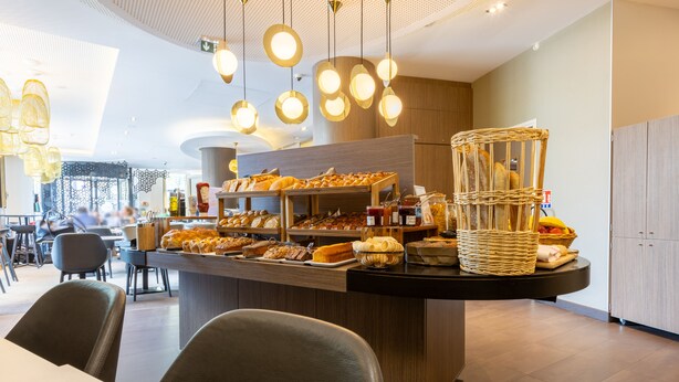 A breakfast bar with various pastries and bread, a wicker basket, and hanging pendant lights in a modern dining area. - Novotel Suites Paris Expo Porte de Versailles (Paris)