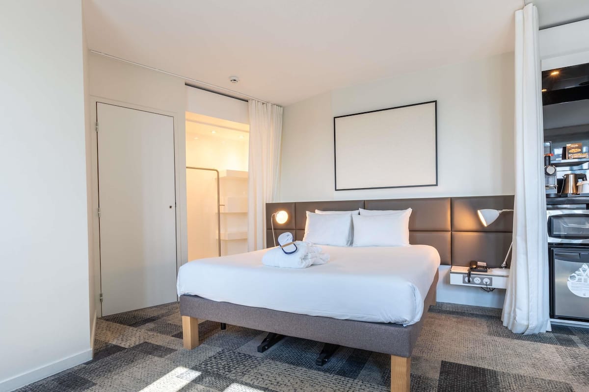 A hotel room with a bed, bedside table, lamp, and a view of the kitchen area.