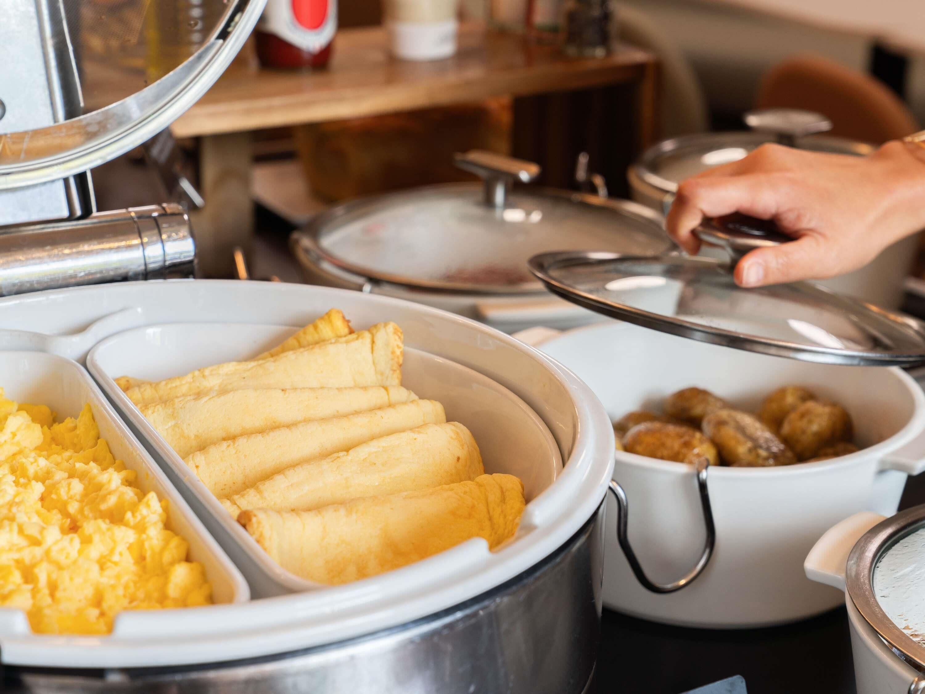A buffet setup with various food items including a bowl of scrambled eggs, a container of fried rolls, and a pot of meatballs.