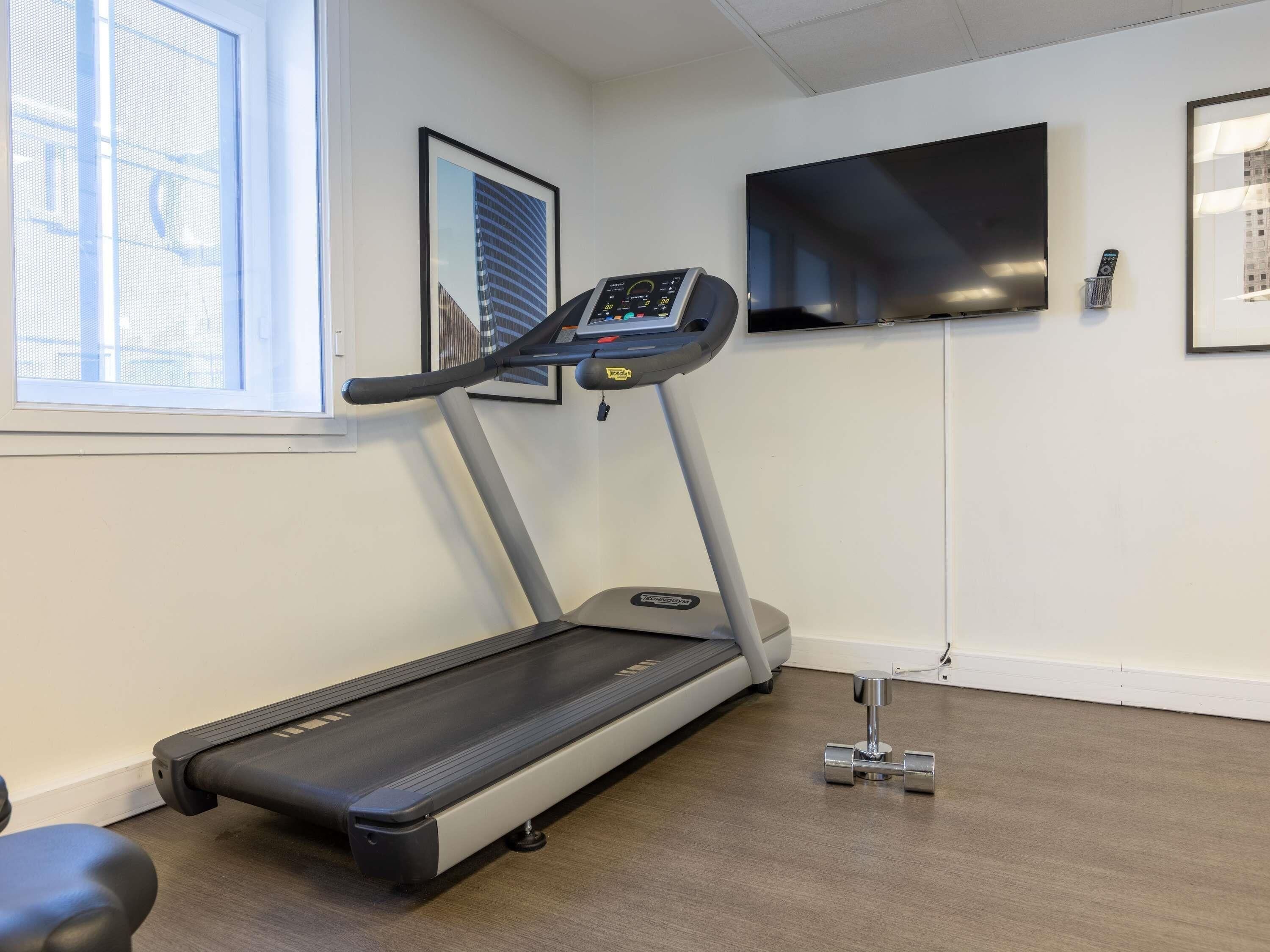 A treadmill in a room with a window, a wall-mounted TV, and a framed picture.