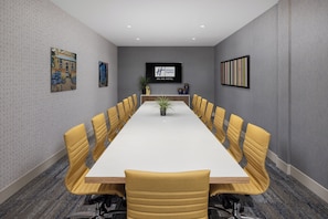 A conference room with a long table, yellow chairs, a potted plant, and a TV screen displaying a logo.