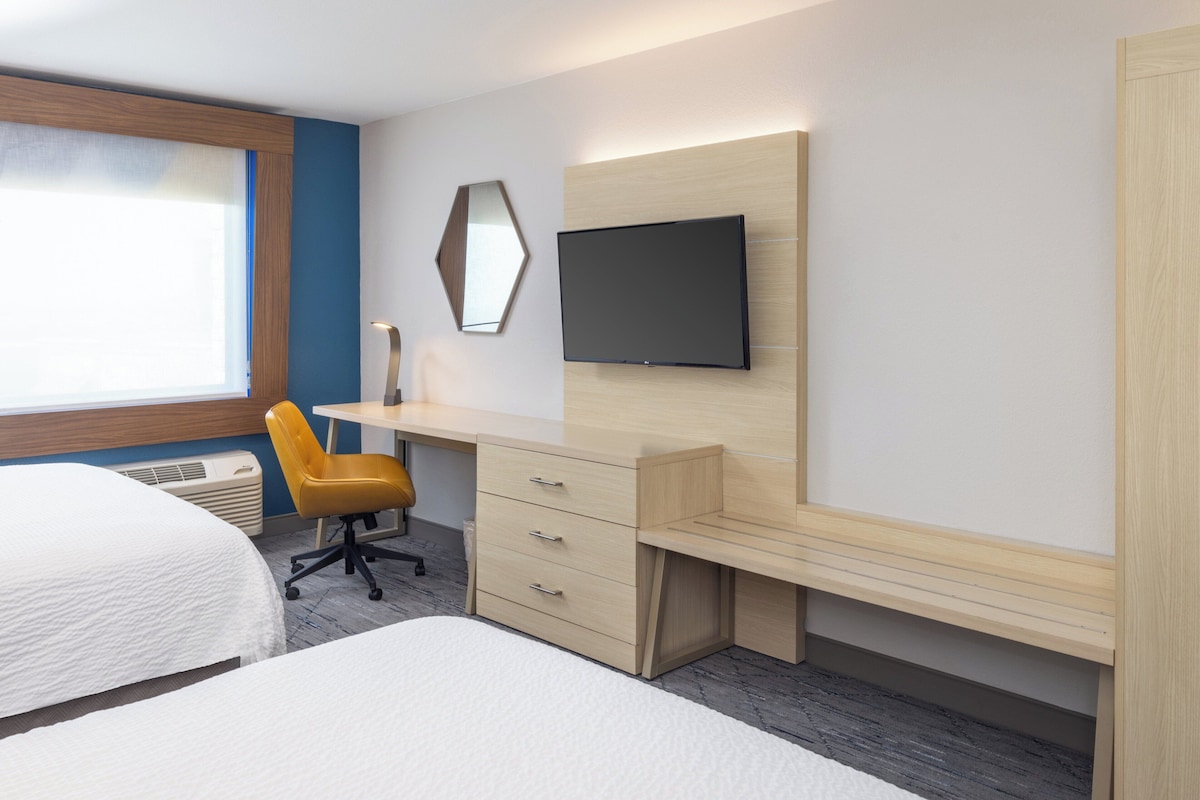 A hotel room with a bed, a desk with a chair, a flat-screen TV, and a window with blinds.