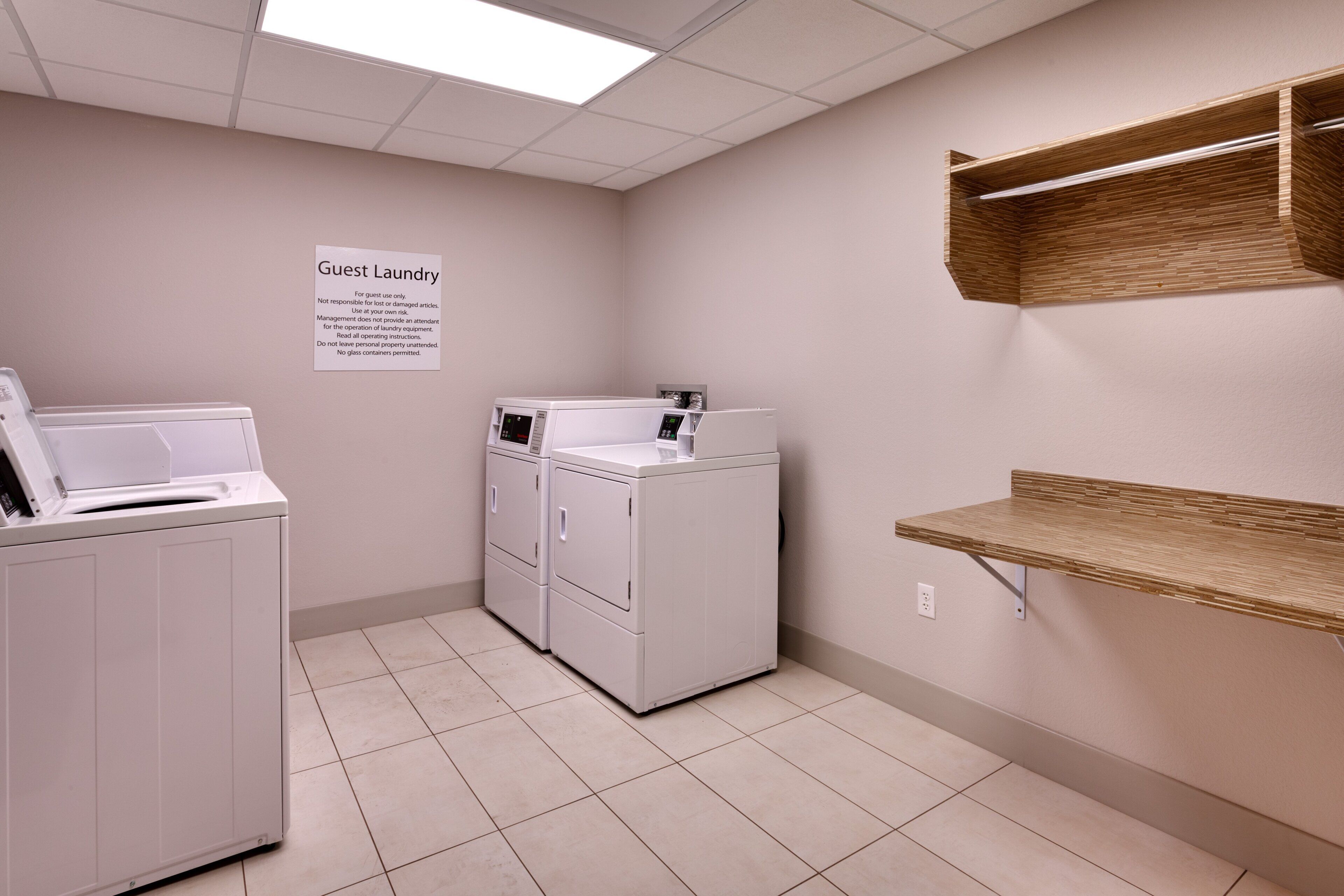 Une buanderie avec deux machines à laver blanches, un panneau indiquant qu’elle est réservée à l’usage des clients, et une étagère avec un panier.