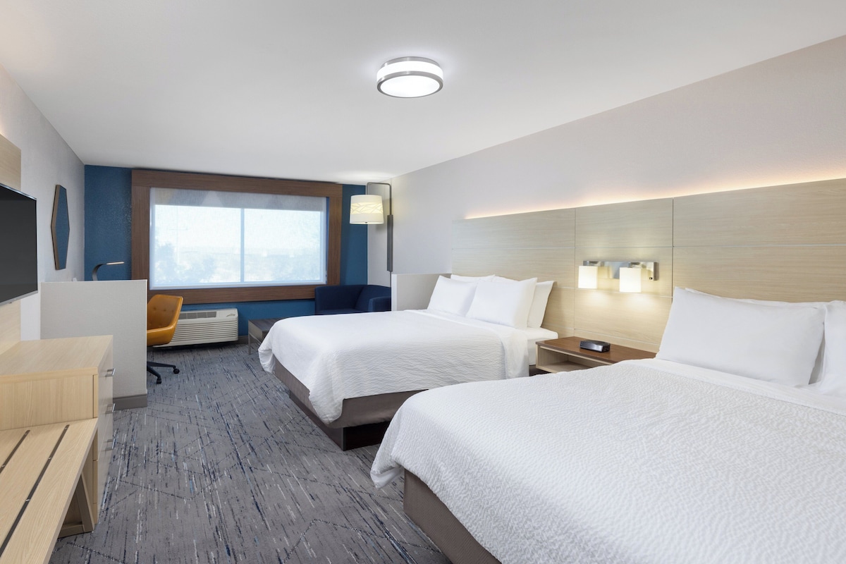 A hotel room with two beds, a desk, a chair, a window, and a wall-mounted lamp.