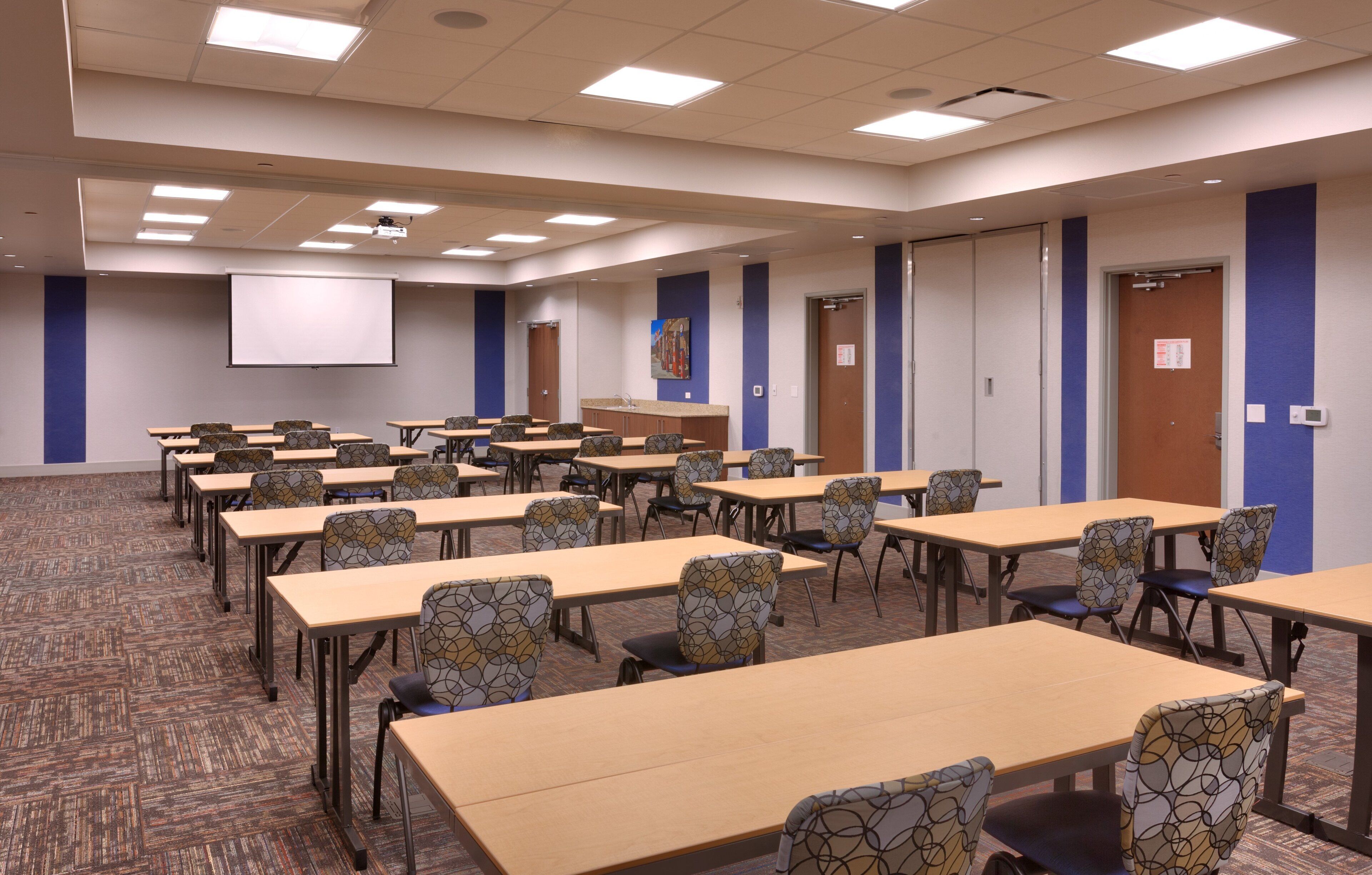 A conference room with multiple tables and chairs, a projector screen, and a carpeted floor.