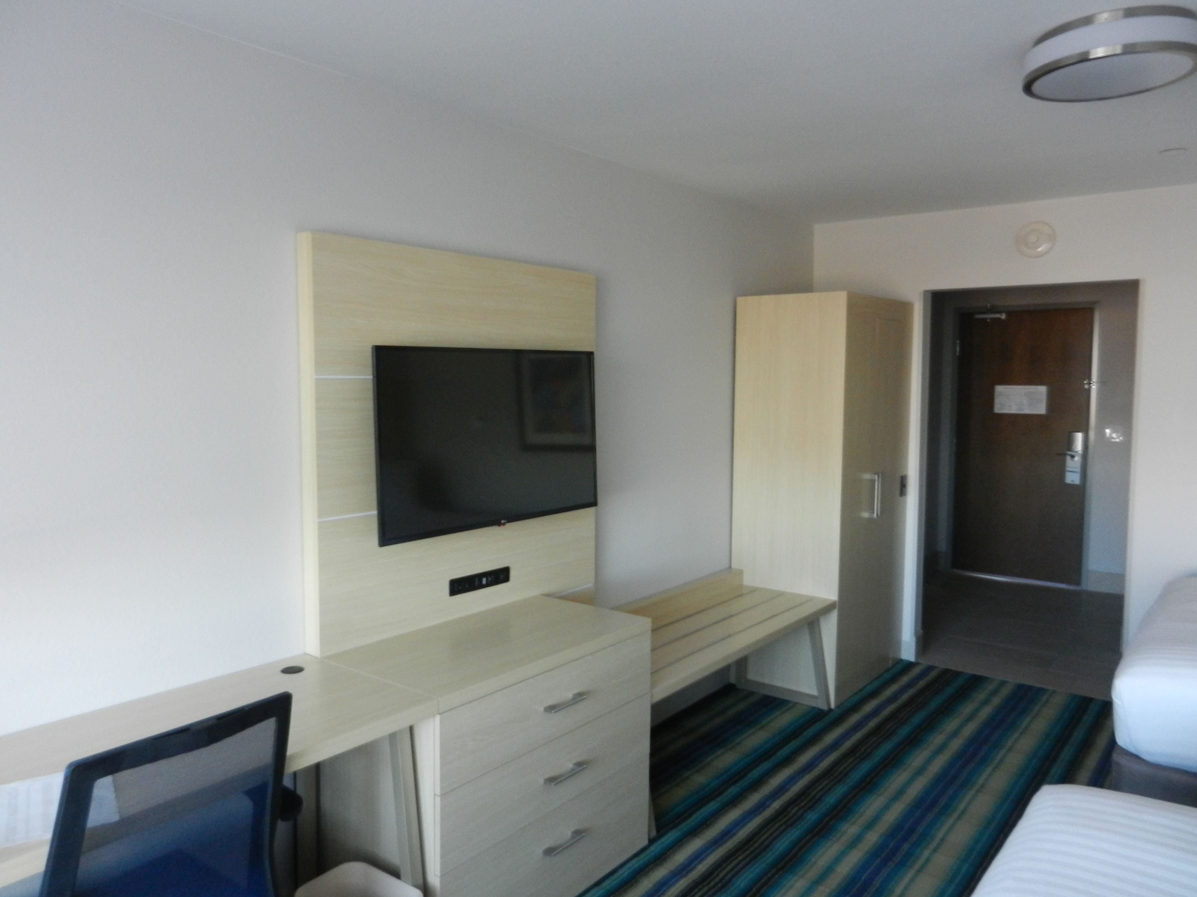 A hotel room with a bed, a desk with a chair, a television mounted on the wall, and a door leading to another room.
