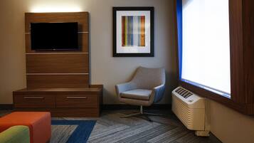A hotel room with a flat-screen TV, a framed abstract painting, a comfortable armchair, a small orange ottoman, and a window with a view.