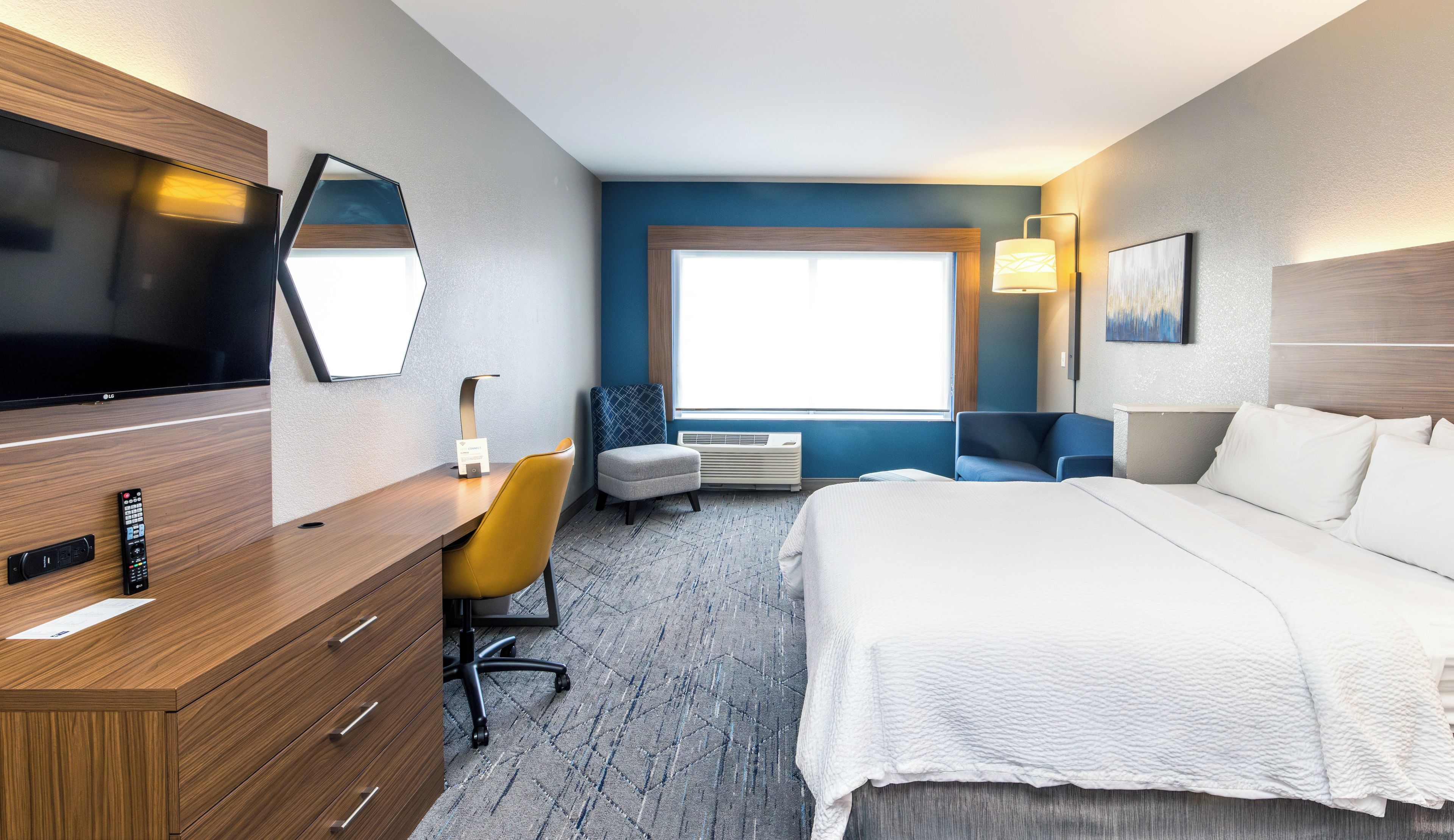 A hotel room with a bed, a desk with a TV, a chair, a window, and a wall-mounted lamp.