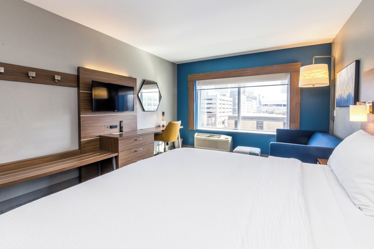 Habitación de hotel con una cama grande, un escritorio con silla, televisión, ventana con vista a edificios y pared azul.