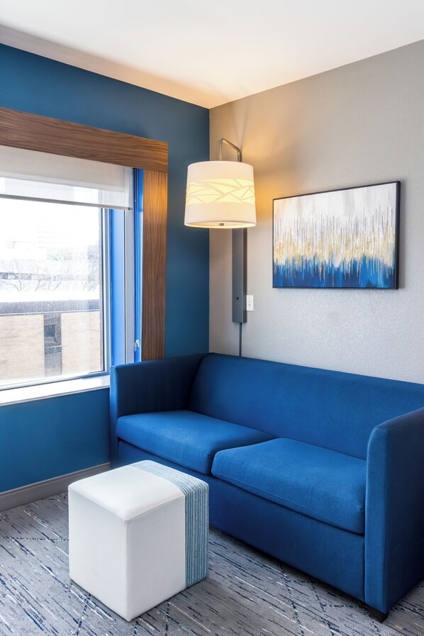 Een moderne kamer met een blauwe bank, een witte poef, een vloerlamp en een stadsgezicht schilderij.