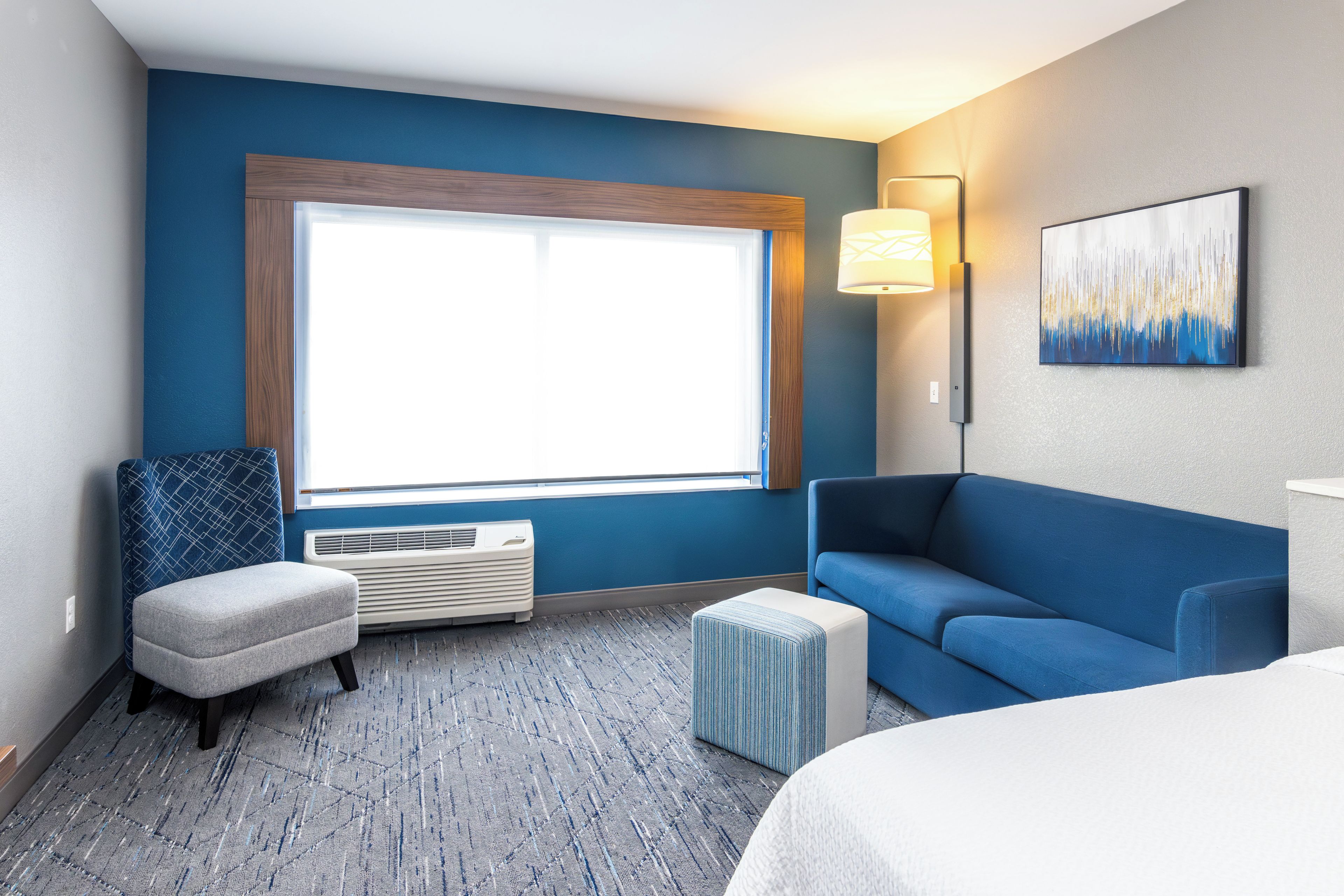 A hotel room with a large window, a blue couch, a small ottoman, a wall-mounted lamp, and a painting.