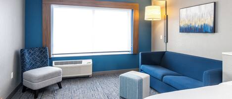 A hotel room with a large window, a blue couch, a small ottoman, a wall-mounted lamp, and a painting.