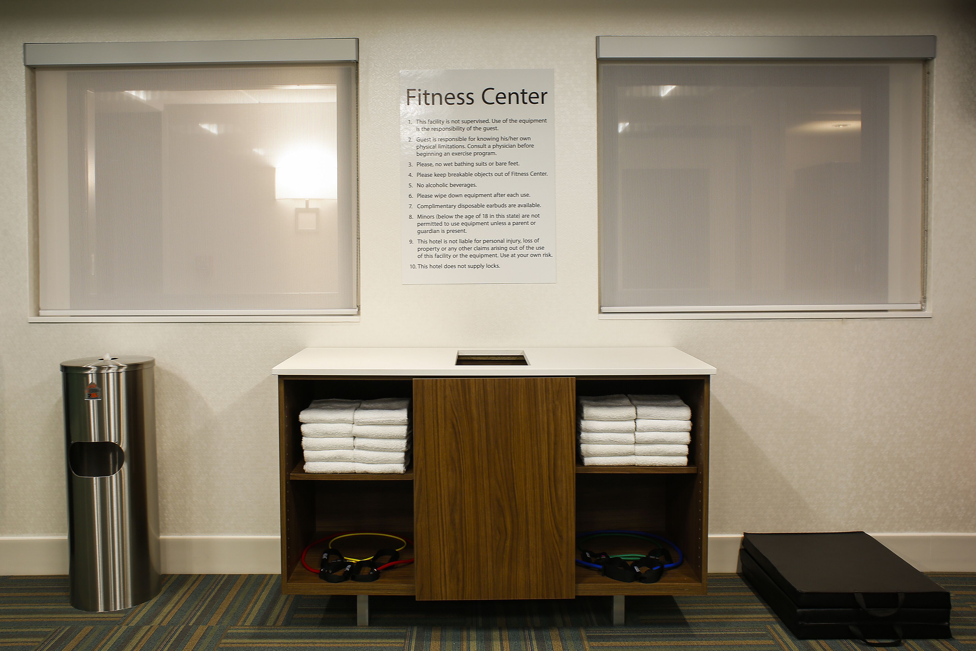 A fitness center with a sign listing rules, a water dispenser, a shelf with towels, and a suitcase.
