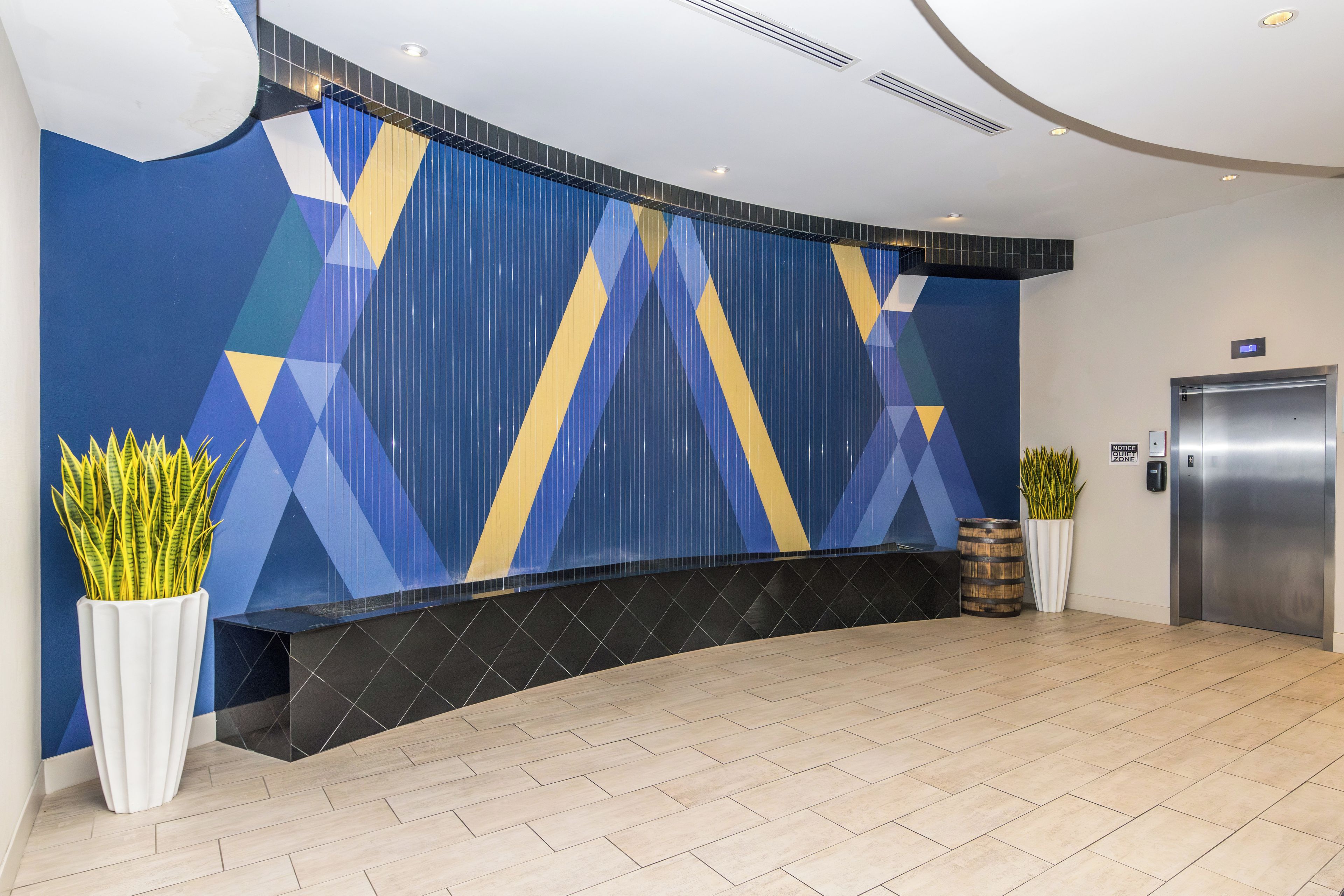 Ett välkomnande område med modern design, med en vägg med geometriskt mönster, en receptionsdisk, en krukväxt och en hiss.