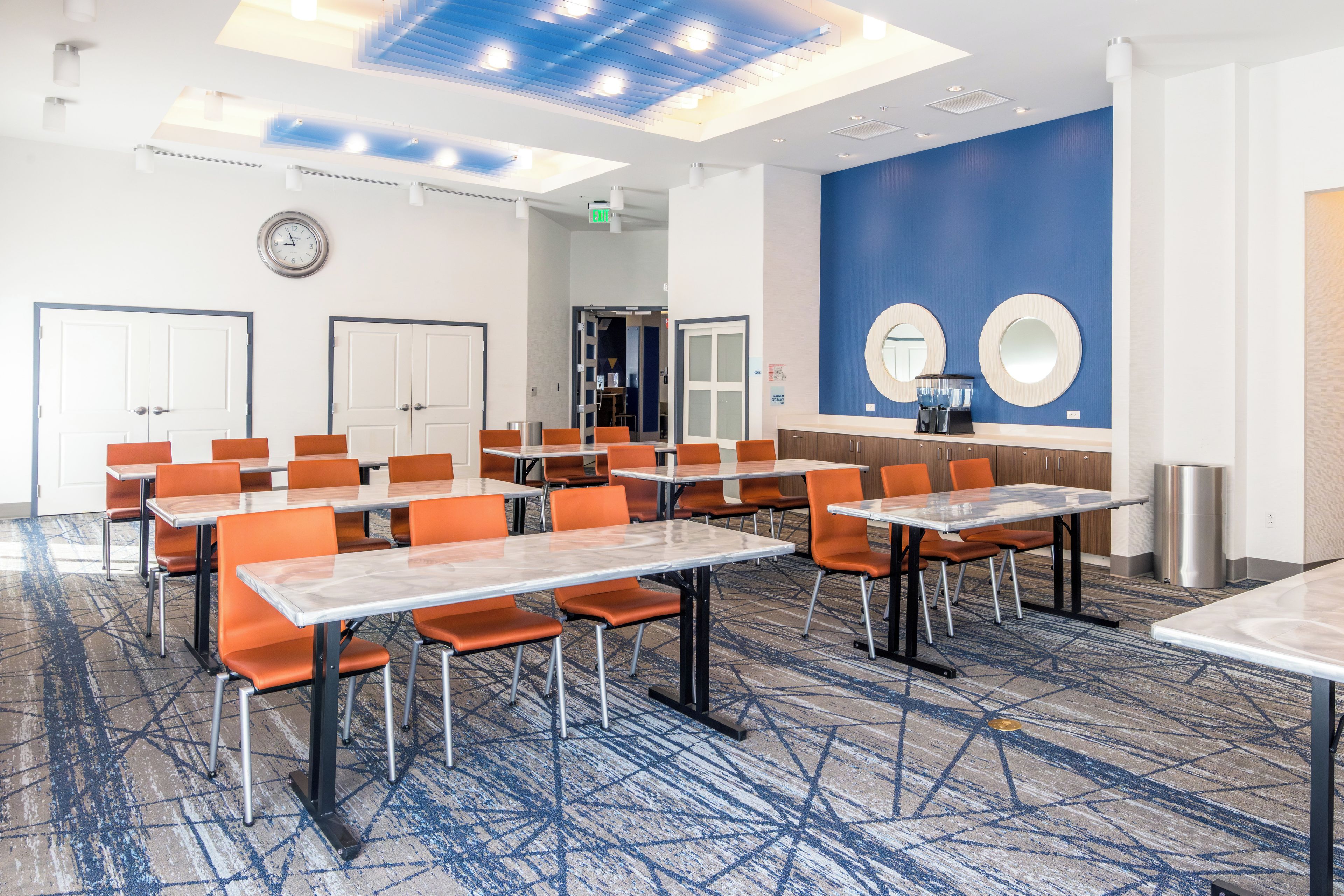 A hotel conference room with multiple tables and chairs, a clock on the wall, and a blue accent wall with two round mirrors.