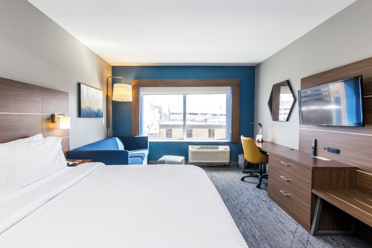 Ein Hotelzimmer mit einem großen Bett, einem Schreibtisch mit Stuhl, einem Fernseher und einem Fenster mit Blick auf die Stadt.