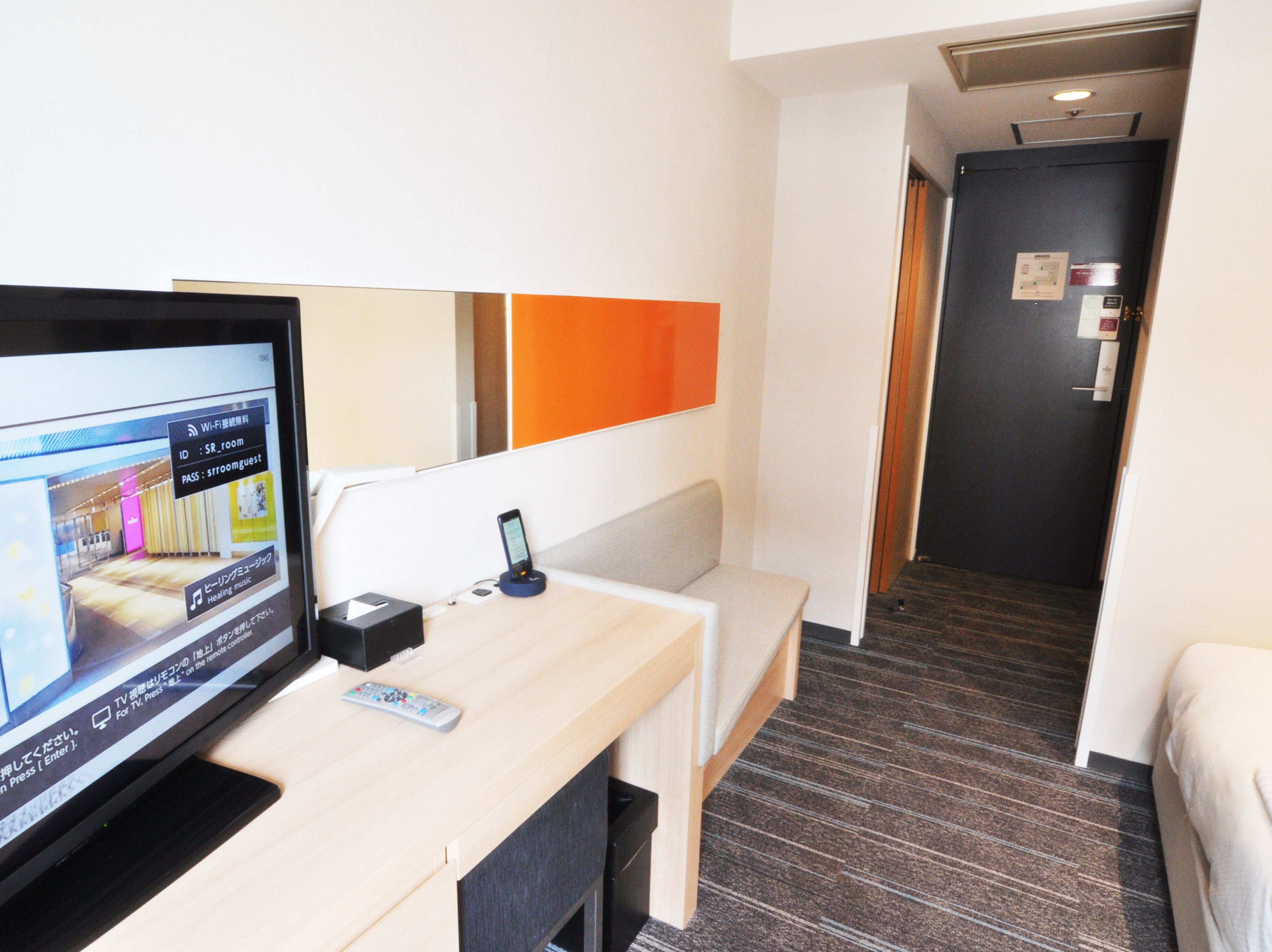 A hotel room with a TV displaying a remote control and a mobile device on a stand, a bed, a desk with a remote control, and a carpeted floor.