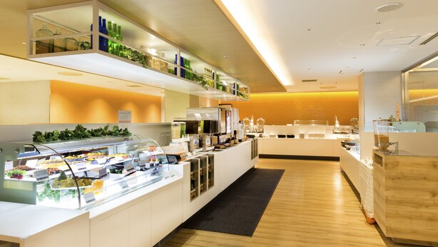 A modern breakfast bar with a glass display case filled with various food items, a coffee station, and a seating area with a wooden floor. - Sotetsu Grand Fresa Osaka - Namba (Osaka)