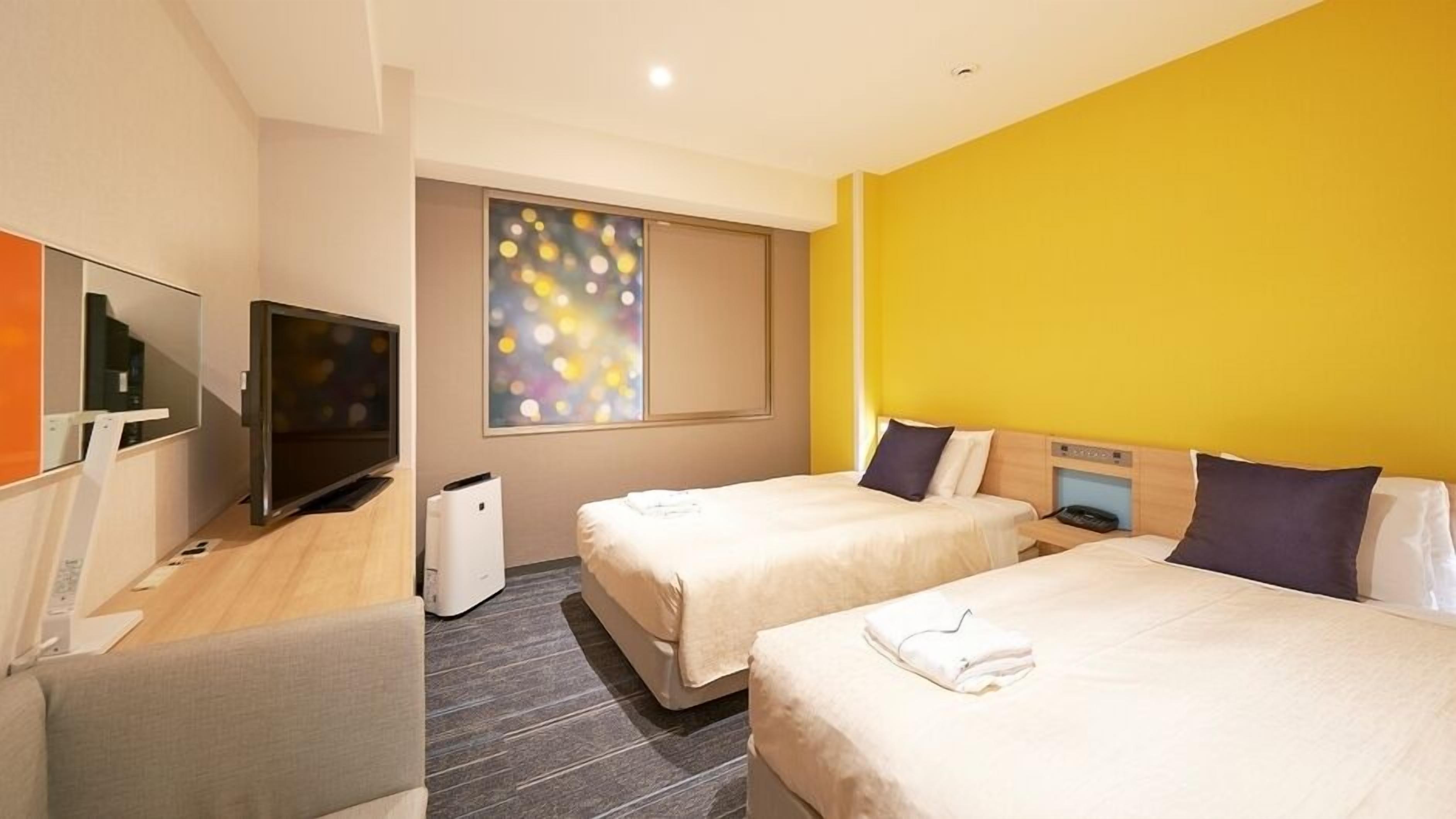 Une chambre d’hôtel avec deux lits, une télévision, une fenêtre donnant sur un beau paysage et un mur orné d’un tableau abstrait aux couleurs vives.
