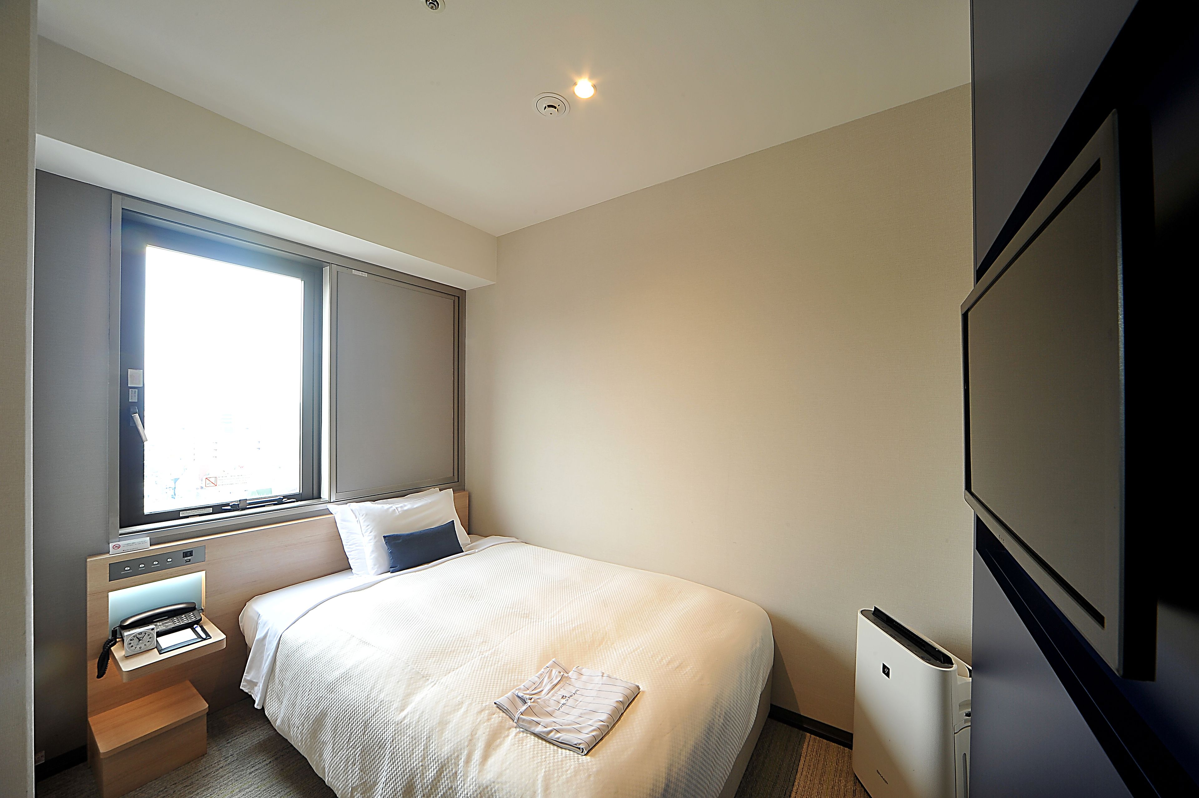 A hotel room with a bed, a bedside table with a phone, a small heater, and a window with blinds.