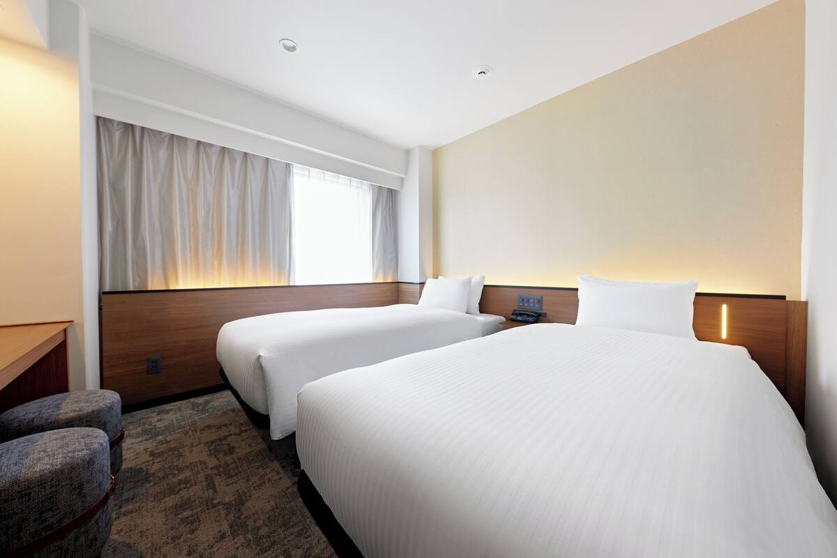 Hotelkamer met twee bedden, een bureau, een stoel, een raam met gordijnen en een aan de muur bevestigde telefoon.