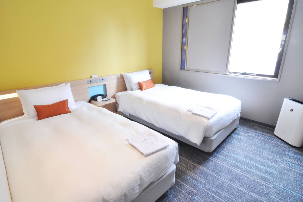 Een hotelkamer met twee bedden, een klein nachtkastje met telefoon, een raam met zonwering en een compact airconditioningapparaat.