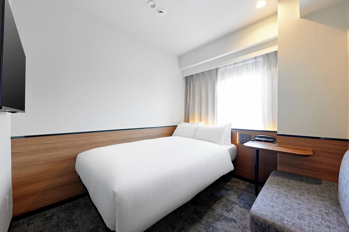 Een hotelkamer met een bed, een klein bureau met telefoon, een stoel, een raam met gordijnen en een flatscreen televisie aan de muur.