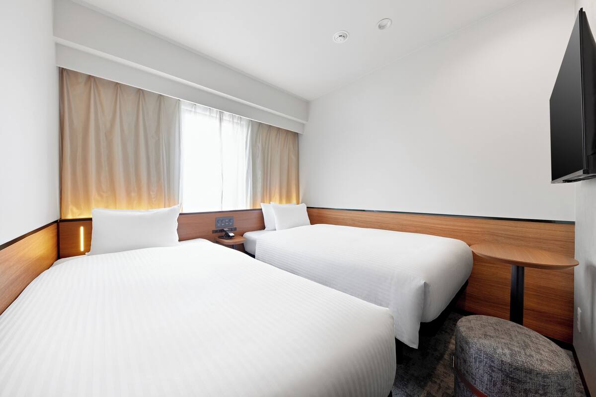 Een hotelkamer met twee bedden, een kleine tafel, een televisie en een raam met gordijnen.