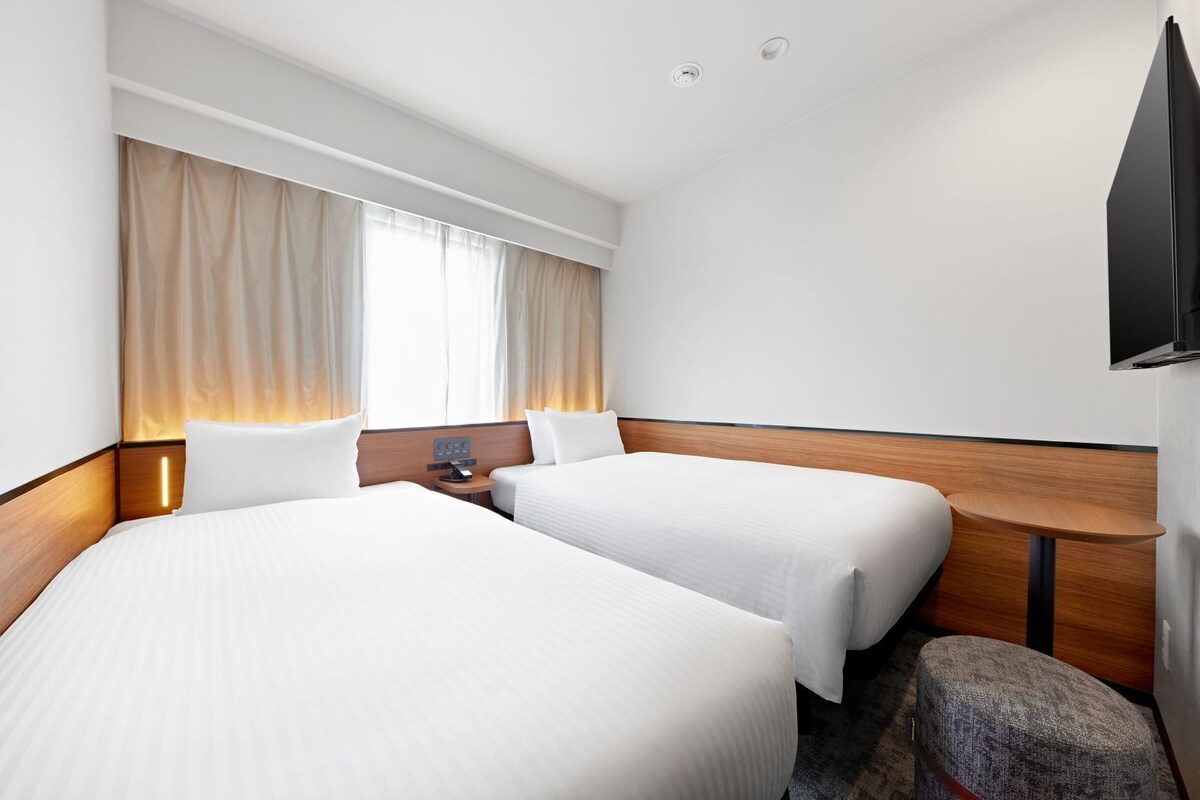 Een hotelkamer met twee bedden, een nachtkastje met lamp, een aan de muur bevestigde televisie en een raam met gordijnen.