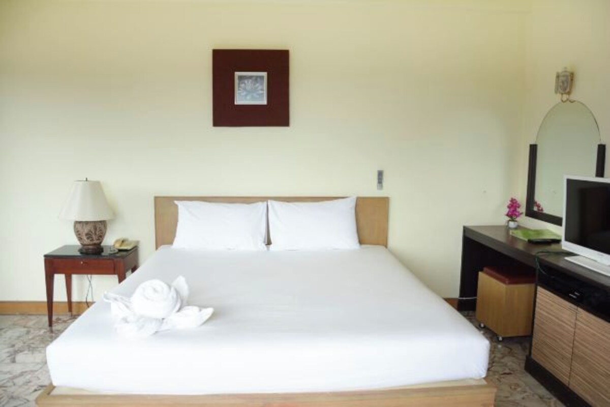 A neatly made bed with white linens, a bedside lamp, a small table with a lamp, a television, and a framed picture on the wall.